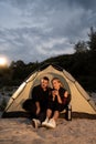 Couple of travelers set up camp on evening lake shore and relaxing in illuminated tourist tent after dinner, drinking