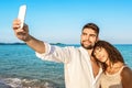 Couple of young lovers taking a selfie on vacation on seashore at sunset or sunrise with boho summer clothes. Afro-American model