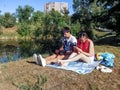A couple of young people with gadgets are sitting on a blanket in a park near the lake. A man with a laptop and a woman with a Royalty Free Stock Photo