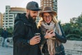 Couple of young travelers are standing on city street and using smartphone.Girl shows guy an image on smartphone screen.
