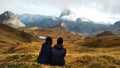 Couple of young travellers sitting on the grass looking to a valley with high mountains, lakes and clouds Royalty Free Stock Photo