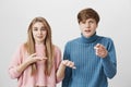 A couple of youngsters pointing with index fingers. Young fair-haired male pointing at camera with dissatisfied face