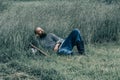 Courageous man with beard resting in the grass in field. Pensive gaze directed to sky. Next to him is ax and warrior helmet