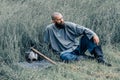 Courageous man with beard resting in the grass in field. Pensive gaze directed to sky. Next to him is ax and warrior helmet