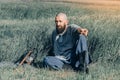 Courageous man with beard resting in the grass in field. Pensive gaze. Next to him is ax and warrior helmet
