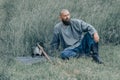 Courageous man with beard resting in the grass in field. Pensive gaze. Next to him is ax and warrior`s helmet