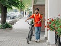 Courier with beard, helmet and yellow bag, walks with bicycle down the street