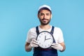 Courier delivery on time! Portrait of positive bearded handyman in overalls and gloves holding clock, smiling to camera