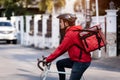 Courier in red uniform with a delivery box on back riding a bicycle and looking on the cellphone to check the address to deliver