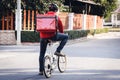 Courier in red uniform with a delivery box on back riding a bicycle and looking on the cellphone to check the address to deliver