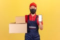 Courier in uniform and protective mask on face holding and showing cardboard boxes and smartphone with empty screen, application Royalty Free Stock Photo
