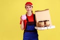 Courier woman holds piggybank and coffee with pizza box, looking at camera with positive expression. Royalty Free Stock Photo