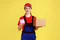 Courier woman standing holding cardboard box and piggybank, looking at camera with happy expression. Royalty Free Stock Photo