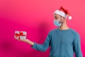 Covid christmas concept. young man with santa claus hat and face mask and gifts in studio with red background, happy smiling face