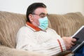 Covid- 19. Elderly man with protective face mask reading a book, while quarantine at home. Royalty Free Stock Photo