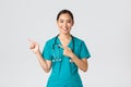 Covid-19, healthcare workers, pandemic concept. Smiling pretty asian doctor, nurse in scrubs and stethoscope, pointing Royalty Free Stock Photo