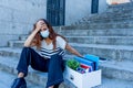 Sad woman in face mask with office box feeling hopeless after losing job due to coronavirus job cuts