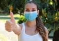 COVID-19 Optimistic sporty girl wearing surgical mask on face during pandemic coronavirus disease showing thumbs up in city park
