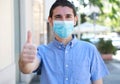 COVID-19 Optimistic young man wearing surgical mask on face during pandemic coronavirus disease showing thumbs up in city street
