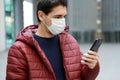 COVID-19 Portrait of young man wearing protective mask reading news on smartphone app in modern city street Royalty Free Stock Photo