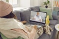 Sick woman wrapped in warm plaids sitting on sofa and talking to online doctor via video call