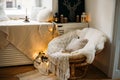 Coziness, comfort, interior and holidays concept - cozy bedroom with chair and christmas garland lights at home. Candle