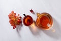 Cozy autumn atmosphere with herbal rose hip tea in transparent teapot with cup, oranges leaves in sun beams on white wood table. Royalty Free Stock Photo