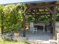 Cozy brown tiber wooden gazebo, pergola with white table, chairs and bench with pillows and blanket surrounded by Royalty Free Stock Photo