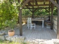 Cozy brown tiber wooden gazebo, pergola with white table, chairs and bench with pillows and blanket surrounded by Royalty Free Stock Photo