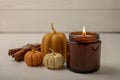 Cozy burning candle in brown glass jar with cinnamon sticks and pumpkin candles on white wood texture. Royalty Free Stock Photo