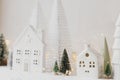Cozy christmas miniature village. Stylish little ceramic houses and trees on snow blanket with golden lights on white background. Royalty Free Stock Photo