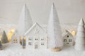 Cozy christmas miniature village. Stylish little ceramic houses and trees on snow blanket with golden lights on white background. Royalty Free Stock Photo