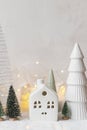 Cozy christmas miniature village. Stylish little ceramic houses and trees on snow blanket with golden lights on white background. Royalty Free Stock Photo