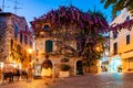 Cozy city streets in Sirmione. Vibrant violet purple flowers growing on facade of medieval building in Old Town. People walking by