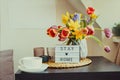 Cozy composition of cup of tea, lightbox with Stay home message and bouquet of fresh spring flowers on the coffee table near