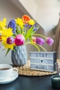 Cozy composition of cup of tea, lightbox with Stay home words and bouquet of fresh spring flowers on the coffee table near window Royalty Free Stock Photo