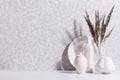 Cozy home decoration with white ceramic vase, pot, dish and bouquet of grey dried herb in morning light on white wood table.