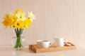 Cozy home interior. Delicate yellow daffodils in glass vase and cup of coffee with milk on a wooden tray on the table. Copy space