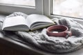 Cozy home still life: cup of hot coffee and opened book with warm plaid on windowsill against snow landscape outside