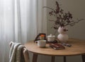 Cozy interior of the living room - cranberry branches in a vase, a lit candle, a cup of tea, a notebook on a round wooden table