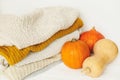 Cozy knitted sweaters and pumpkins on white background. Hello fall! Stylish white and yellow sweaters stack and orange pumpkins