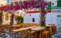 Cozy outdoor dining, cafe, restaurant, taverna in small Greek square, whitewashed chapel, vibrant pink bougainvillea in Royalty Free Stock Photo