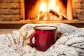 Cozy scene near fireplace with a Red enameled mug with hot tea and cozy warm scarf. Royalty Free Stock Photo