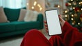 Cozy setting with person holding smartphone featuring blank white screen, surrounded by festive decorations and Royalty Free Stock Photo