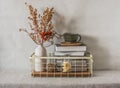 Cozy simple home interior - books in a metal basket, a ceramic cup, dried flowers in a vase on the table. Cozy home interior