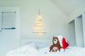 Cozy toy bear in santa hat sitting on the bed on background of Unusual Christmas tree made of wooden sticks with a garland on the