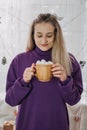 Cozy winter Christmas mood. Young woman at home dress with coffee Cup and marshmallows in hand standing near xmas tree Royalty Free Stock Photo