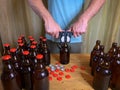 Craft beer brewing at home, man closes brown glass beer bottles with plastic capper on wooden table with red crown caps.