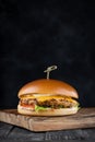 Craft juicy cheeseburger served with a wooden skewer on a wooden cutting board on a smoky black background with