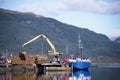 Crane lifting wood logs from stack to ship in sea loch for transportation of biomass fuel in Scotland